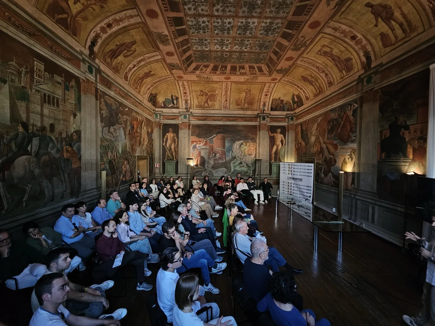 La sala affrescata dell'Humanitas a Ferrara piena di persone sedute, anche a terra